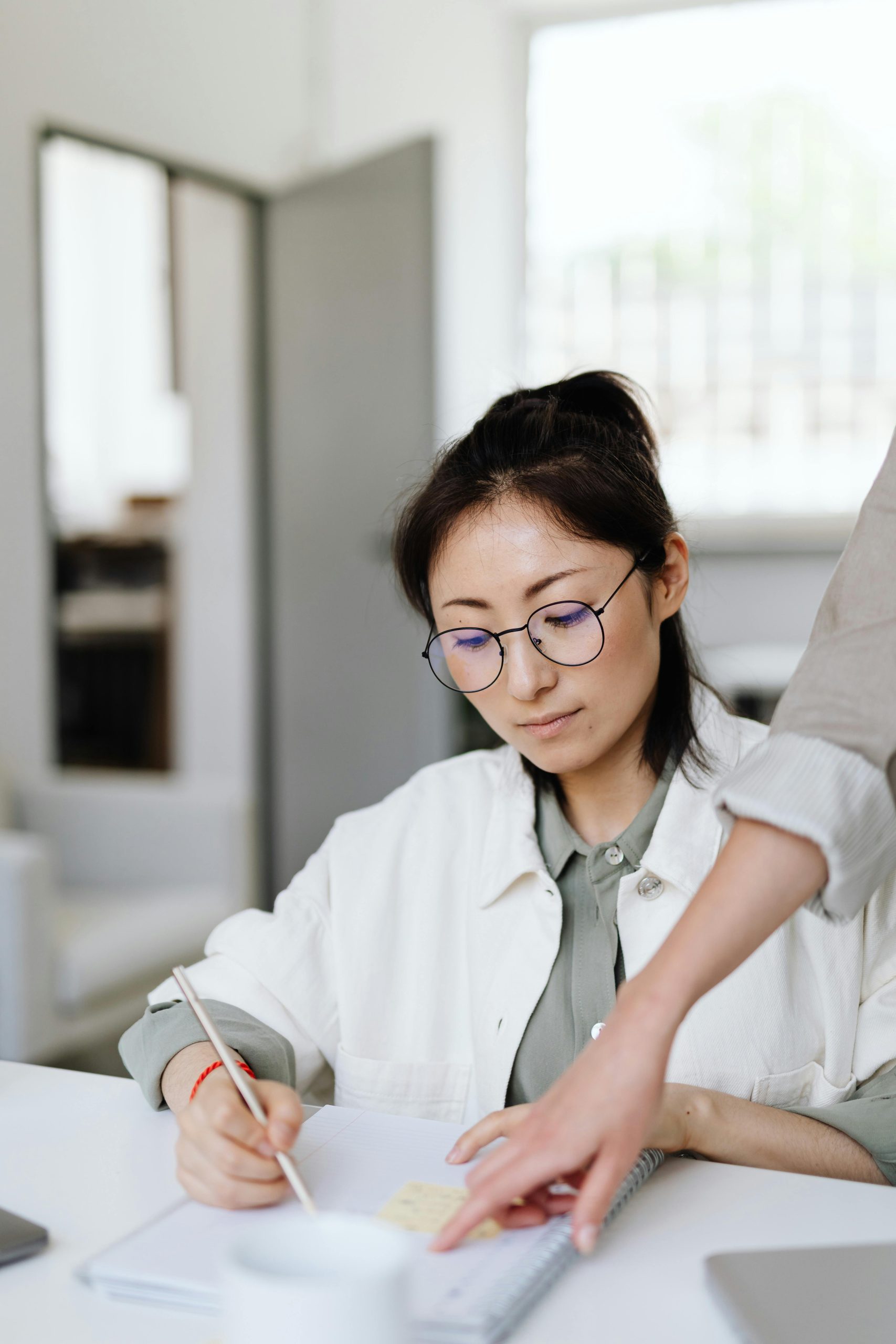 Young woman with glasses studying at a desk, guided by an instructor inside a classroom.